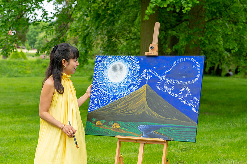 Lorraine Artsy standing beside her original acrylic painting inspired by Mayon Volcano in the Philippines, displayed outdoors in a peaceful green park setting under a bright full moon design.